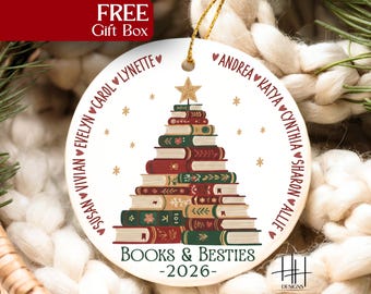 May include: A round white ceramic Christmas ornament featuring a stack of books arranged in a tree shape topped with a gold star. The text reads "BOOKS & BESTIES -2026-" with a list of names separated by hearts encircling the book tree design.