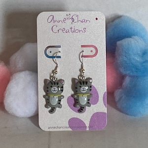 May include: Dangle earrings with grey cat charms, black stripes, and yellow collars. The earrings hang from silver hooks. The earrings are displayed on a card with the text "Anne Chan Creations".
