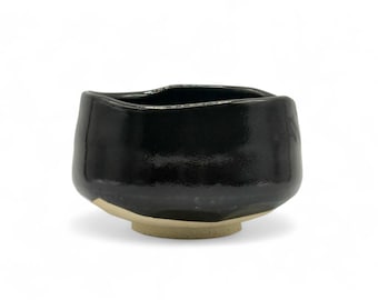 May include: A black ceramic tea bowl with a slightly irregular shape and a light brown rim. The bowl is designed for whisking matcha tea.