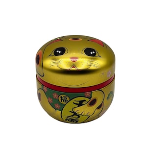 May include: Gold-colored Maneki Neko cat-shaped container. The container features a smiling cat face on the top and a waving paw design on the side. The cat has black eyes and a pink nose. The container has a red band around the middle.