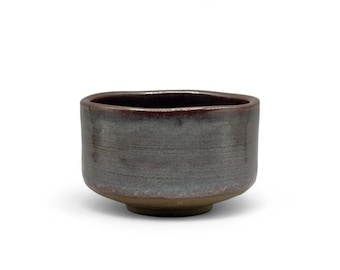 May include: A dark gray ceramic tea bowl with a slightly uneven rim. The bowl has a subtle iridescent glaze.