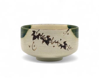 May include: A white ceramic tea bowl with a green rim and brown leaf design. The bowl is a traditional Japanese tea ceremony bowl.