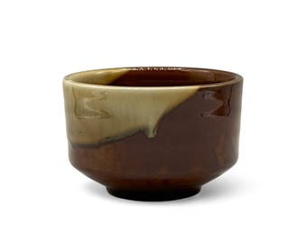 May include: A small, handcrafted ceramic bowl with a rounded shape. The bowl features a two-tone glaze, with a light beige interior and a rich, dark brown exterior. The bowl is designed for serving or display.