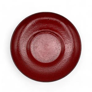 May include: A deep red ceramic saucer with a slightly raised center. The saucer has a textured surface and a glossy finish.
