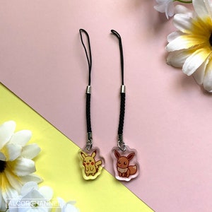 Puede incluir: Dos charms de teléfono negros con charms de plástico transparente que presentan imágenes de dibujos animados de Pikachu y Eevee. Los charms cuelgan de cordones trenzados negros.