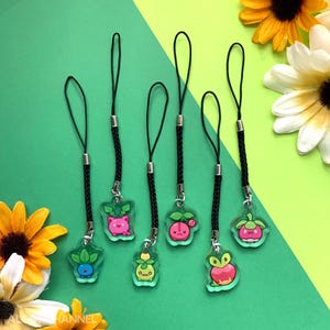 Puede incluir: Seis colgantes de acrílico transparente con diseños de plantas de dibujos animados. Cada colgante tiene un cordón negro y un cierre de metal plateado. Los colgantes presentan colores rosa, verde y rojo, sobre un fondo verde y amarillo.