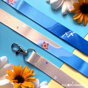 May include: Three lanyards with beach-themed designs and a silver-colored clasp. One lanyard is beige with a starfish pattern, another is blue with a wave pattern, and the third is blue with a sun and wave pattern. The lanyards are on a blue background with flowers.