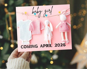 May include: A pink card with the words "baby girl" and "COMING APRIL 2026". The card features a baby onesie, a sweater, and two bunny dolls. The background is a blurred Christmas tree.
