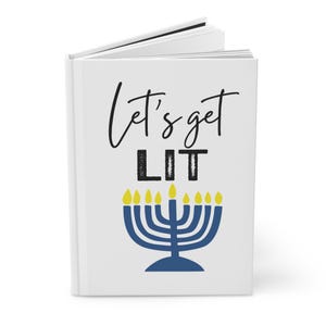 Puede incluir: Diario blanco con el texto "Let's get LIT" en escritura negra y letras de imprenta. Una menorá azul con llamas de velas amarillas aparece debajo del texto. El diario está abierto, revelando páginas en blanco.