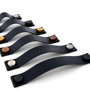 May include: A set of six black leather cabinet pulls with different coloured metal accents. The pulls are arranged in a row, with the metal accents facing the viewer. The pulls are all the same size and shape, and they have a simple, modern design.