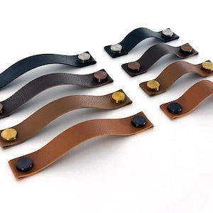May include: Eight leather cabinet pulls with metal accents in various colors, including brown, black, and gold. The pulls are curved and have a simple design.