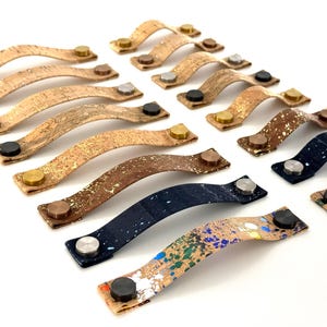 May include: A collection of decorative cabinet pulls with a curved design. The pulls are made of cork and feature various finishes, including natural cork, metallic accents, and paint splatters. Each pull has a metal button on each end.
