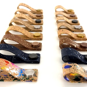 May include: A set of 18 cork and metal strap loops in various colours and patterns, including natural cork, brown, blue, and multi-coloured. The straps have a loop on one end and a metal button on the other.