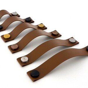 May include: Six brown leather cabinet pulls with various metal rivet colors: silver, black, gold, copper. The pulls have a curved design and are set against a white background. These are ideal for furniture upgrades.