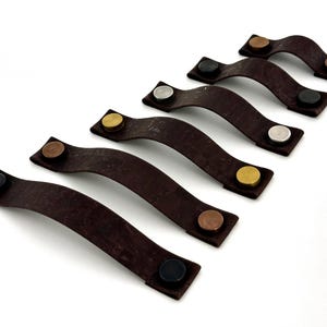 Peut inclure: Six poignées de tiroir en cuir brun avec des accents métalliques de différentes couleurs. Les poignées sont disposées en rangée du plus long au plus court.