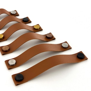 May include: Six brown leather cabinet pulls with different colored metal accents. The pulls are arranged in a row, with the top pull being the shortest and the bottom pull being the longest.