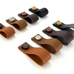 May include: Assortment of eight leather cabinet pulls with different colored metal hardware. The pulls are brown, black, and tan.