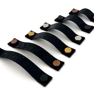May include: Six black leather cabinet pulls with different colored metal accents. The pulls are arranged in a row from shortest to longest.