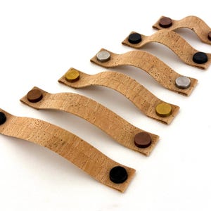 Può includere: Un set di sei tiretti per cassetti in sughero con pomelli in metallo di diversi colori. I tiretti sono realizzati in sughero naturale e hanno una forma curva. I pomelli sono di colore nero, argento, oro, marrone e rame.