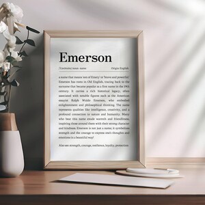 Peut inclure: Impression encadr&eacute;e avec le nom "Emerson" et sa d&eacute;finition. Un vase avec des fleurs blanches est &agrave; gauche. L'impression est sur une surface en bois. Le texte est en police serif.