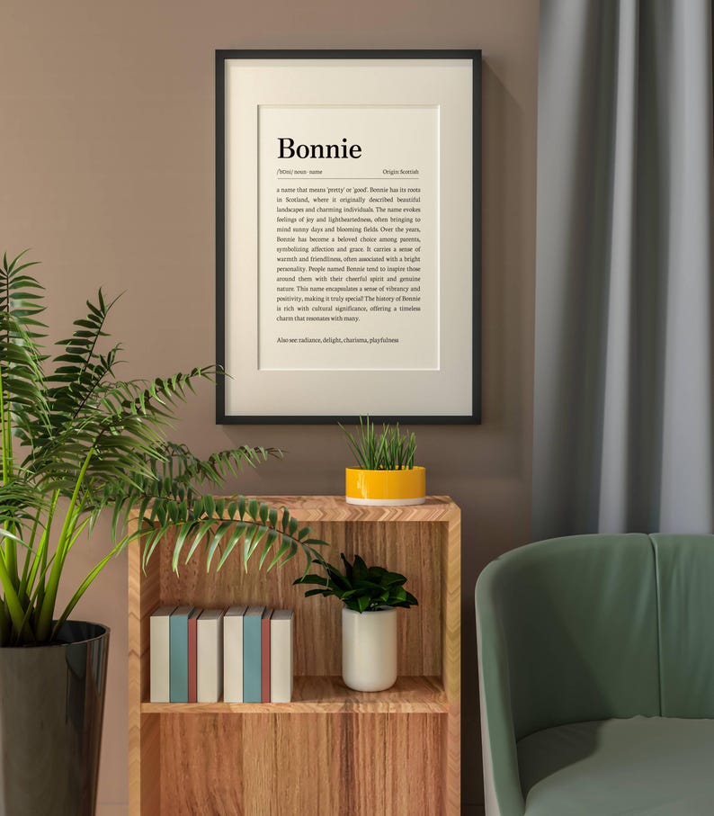 May include: A framed print with the name "Bonnie" in a black frame. The print features a definition of the name, with text in a serif font. The print is displayed on a wall above a wooden shelf with plants and books.