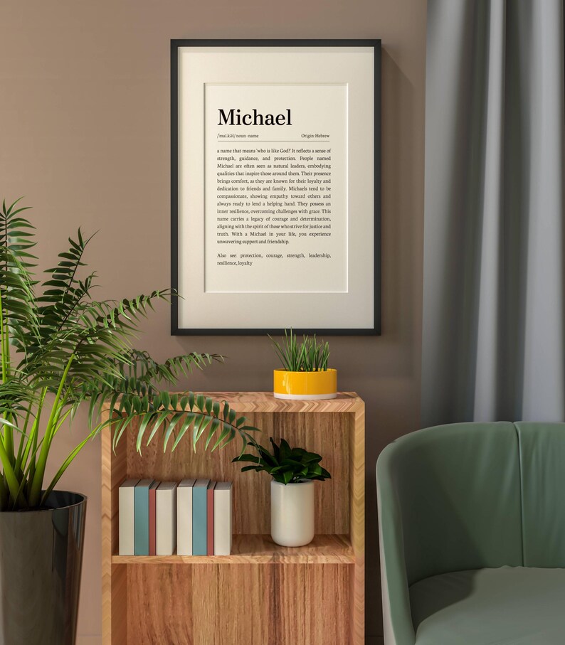 May include: A framed print with the name "Michael" in a black frame. The print includes a definition of the name, its origin, and associated qualities. The print is displayed on a wall above a wooden shelf with books and plants.