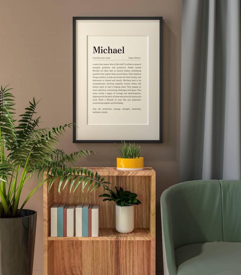 May include: A framed print with the name "Michael" in a black frame. The print includes a definition of the name, its origin, and associated qualities. The print is displayed on a wall above a wooden shelf with books and plants.