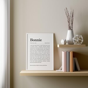 May include: A framed print with the name "Bonnie" in a minimalist style. The print is displayed on a light-colored wooden shelf, alongside decorative items like a white vase with branches, geometric shapes, and books.