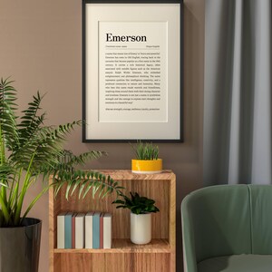 Peut inclure: Un cadre accroch&eacute; au mur avec le nom "Emerson" et sa d&eacute;finition. L'impression est dans un cadre noir et est suspendue au-dessus d'une &eacute;tag&egrave;re en bois avec des livres et des plantes. Une chaise verte et un rideau sont en arri&egrave;re-plan.