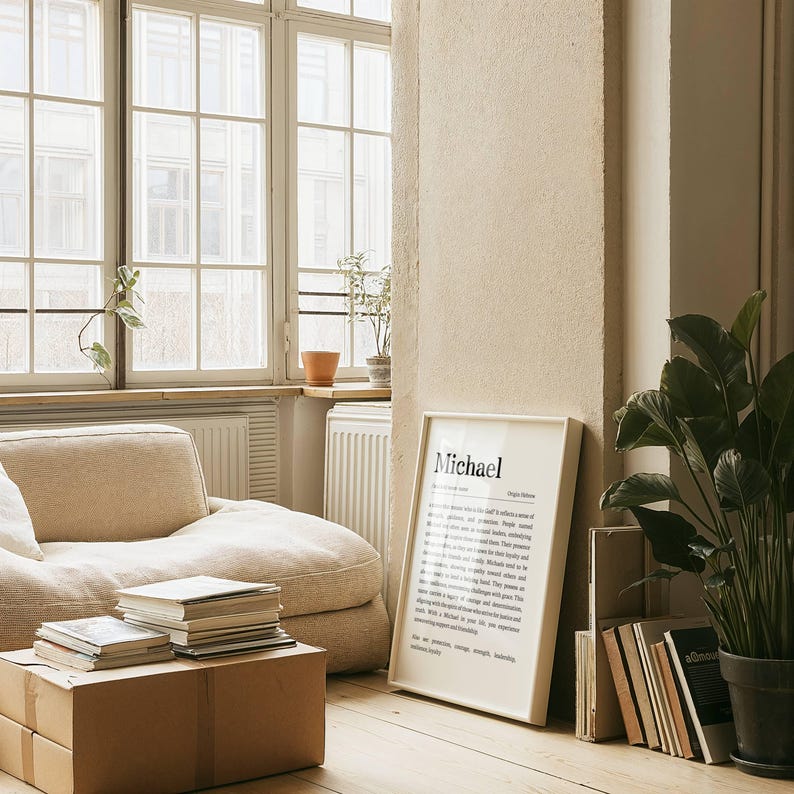 May include: A framed print with the name "Michael" in black text, leaning against a wall. The print is in a room with a beige beanbag chair, a stack of books, and a potted plant. The room has large windows.