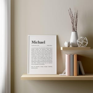 May include: A framed white print with the name "Michael" and its definition. The print is displayed on a light wooden shelf, alongside decorative items like a white vase with branches, geometric shapes, and books.