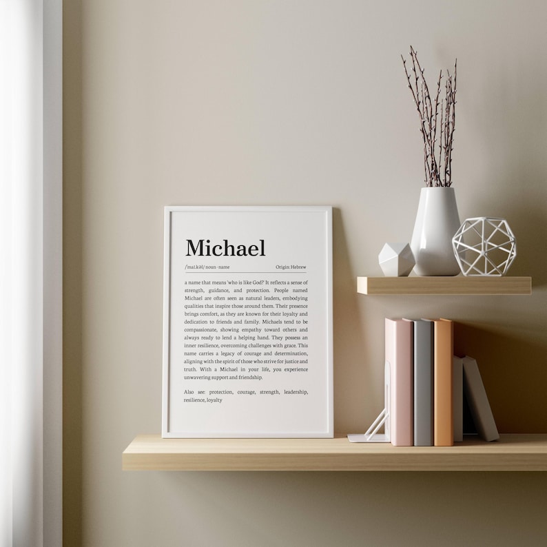 May include: A framed white print with the name "Michael" and its definition. The print is displayed on a light wooden shelf, alongside decorative items like a white vase with branches, geometric shapes, and books.