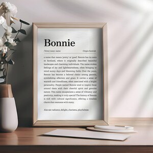 May include: A framed print with the name "Bonnie" in a serif font. The print includes a definition and origin of the name. A vase with white flowers sits to the left of the frame. A pen and paper are on the table.
