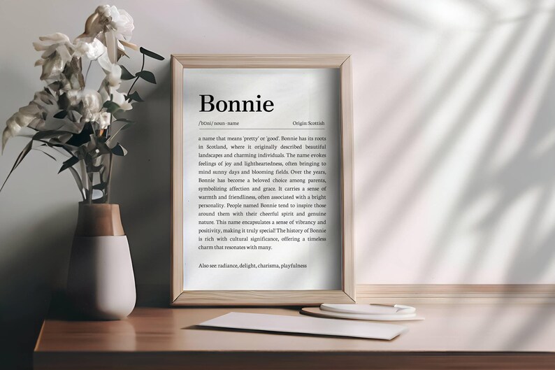 May include: A framed print with the name "Bonnie" in a serif font. The print includes a definition and origin of the name. A vase with white flowers sits to the left of the frame. A pen and paper are on the table.