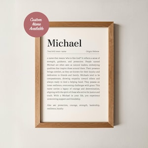 May include: A framed print featuring the name "Michael" in a bold font, with a definition and origin. The print is in a wooden frame and includes a pink circle with the text "Custom Name Available". The text describes the name's meaning and associated qualities.