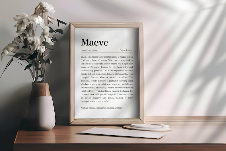 May include: A framed print with the name "Maeve" in a serif font, with a definition and origin. The print is in a light wood frame, resting on a wooden surface. A vase with white flowers sits to the left.