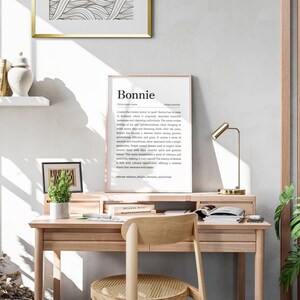 May include: A light wood desk with a framed print that reads "Bonnie" sits in a well-lit room. A small potted plant, a gold desk lamp, and a wooden chair are also visible. A decorative shelf with gold accents is in the background.