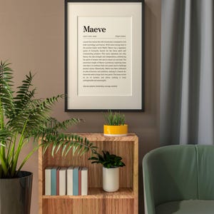 May include: A framed print with the name "Maeve" displayed in a modern font. The print includes a definition and origin information. The frame is black, and the print is displayed on a wall with a wooden shelf and plants.