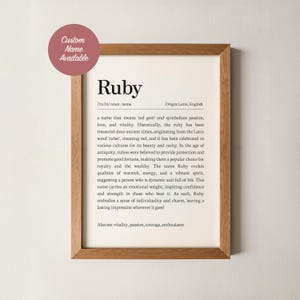 May include: A framed print with the name "Ruby" in a serif font, accompanied by its pronunciation, origin, and a definition. The print is in a wooden frame, and a circular sticker in the upper left corner reads "Custom Name Available."