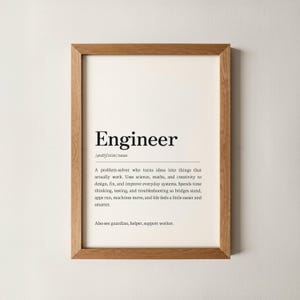 May include: A framed print with the definition of "Engineer" in black text on an off-white background. The definition includes the phonetic pronunciation and a description of an engineer's role. The frame is made of light brown wood.