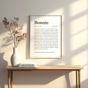May include: A framed print with the name "Bonnie" in a minimalist design. The print includes a definition, origin, and meaning of the name. The print is displayed on a wooden table with a vase of dried flowers and a stack of books.