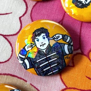 May include: A round, yellow button with a cartoon illustration of a person covering their ears. The person is wearing a black jacket with white stripes and the initials "HG". The button has a holographic effect.