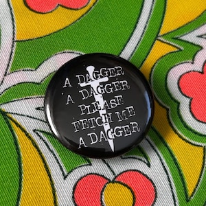 May include: A black button with white text that reads "A DAGGER A DAGGER PLEASE FETCH ME A DAGGER" and a white dagger graphic. The button is set against a colorful floral patterned background.