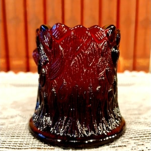 May include: A ruby red glass votive candle holder with a detailed, textured pattern. The candle holder has a flared base and a scalloped rim. The design features a repeating leaf pattern. The candle holder is approximately 3 inches tall.