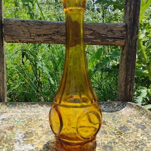 Puede incluir: Un jarrón de vidrio ámbar vintage con un cuello delgado y una base decorativa redondeada. El jarrón presenta diseños circulares y una parte superior acampanada. El jarrón se exhibe sobre una bandeja con estampado floral, con una silla de madera y vegetación en el fondo.