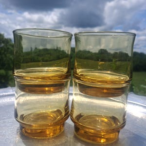 May include: Four amber-colored drinking glasses stacked in pairs. The glasses have a cylindrical shape with a slightly flared rim and a thick base. They are sitting on a silver tray, with a backdrop of a cloudy sky and green foliage.