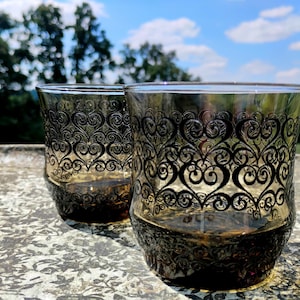 May include: Two clear drinking glasses with a decorative black heart pattern. The glasses are made of a smoky gray glass and are sitting on a stone surface. The background includes trees and a blue sky.