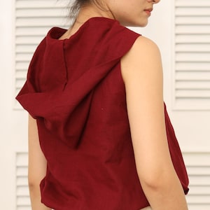 以下が含まれることがあります： 大きなフードが付いた、袖なしのワインレッドのトップス。トップスは柔らかく、流れるような生地で作られています。