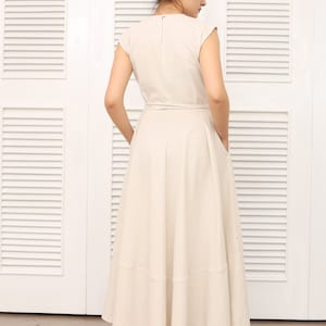 May include: A beige linen dress with a wrap style bodice and a flowing skirt. The dress has a simple, elegant design and is perfect for a casual summer occasion.