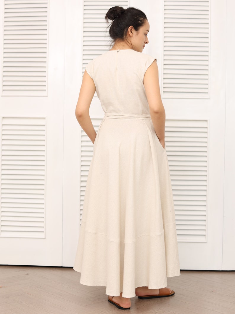 May include: A beige linen dress with a wrap style bodice and a flowing skirt. The dress has a simple, elegant design and is perfect for a casual summer occasion.